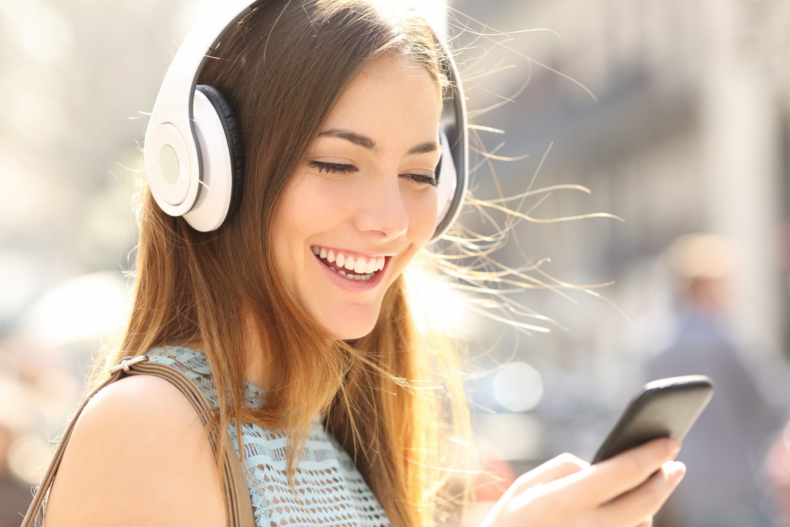 Portrait of a happy girl listening music on line with wireless headphones from a smartphone in the street in a summer sunny day