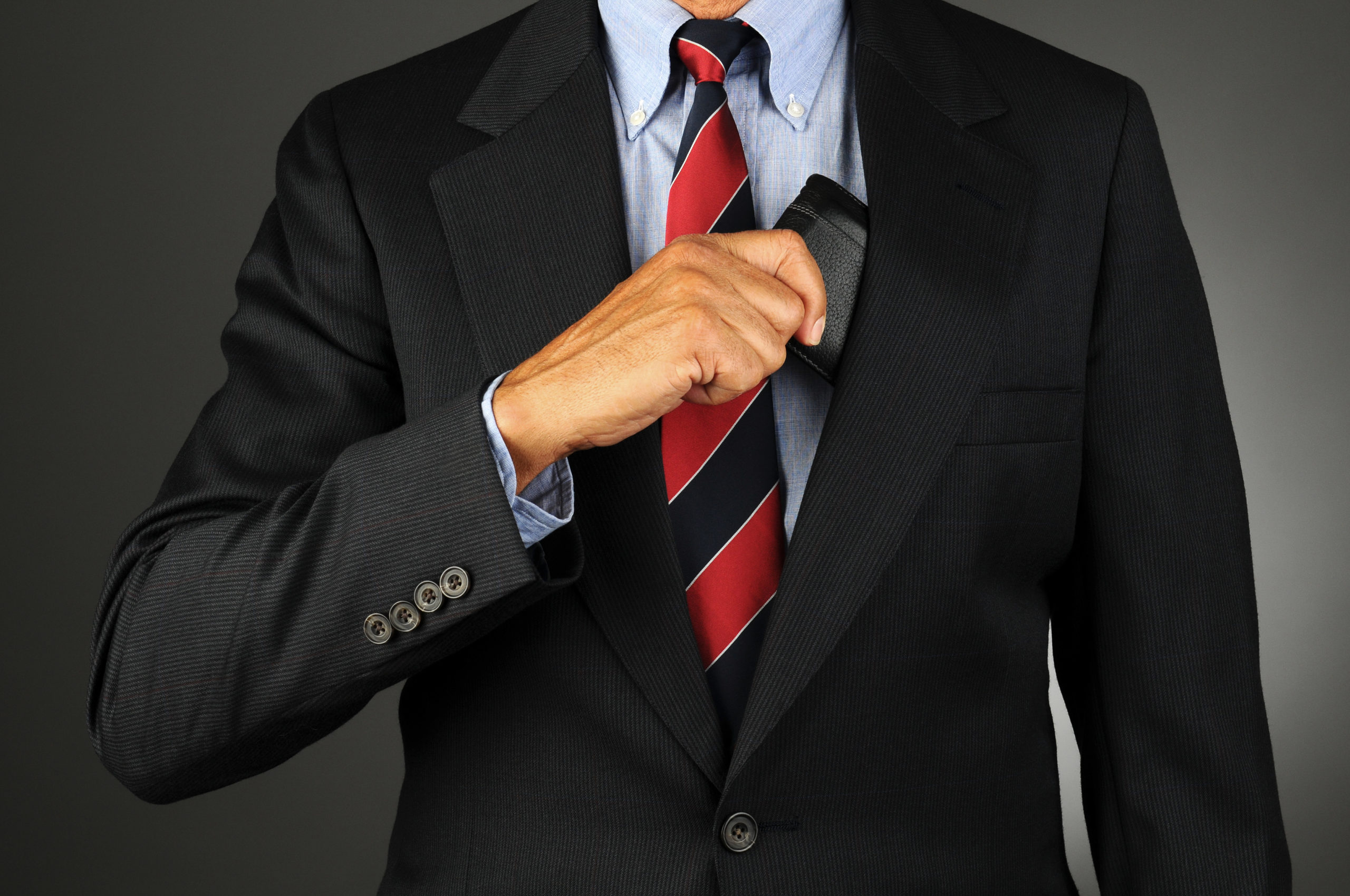 Closeup of a businessman reaching into his coat pocket to get his wallet. Horizontal format over a light to dark gray background. Man is unrecognizable.